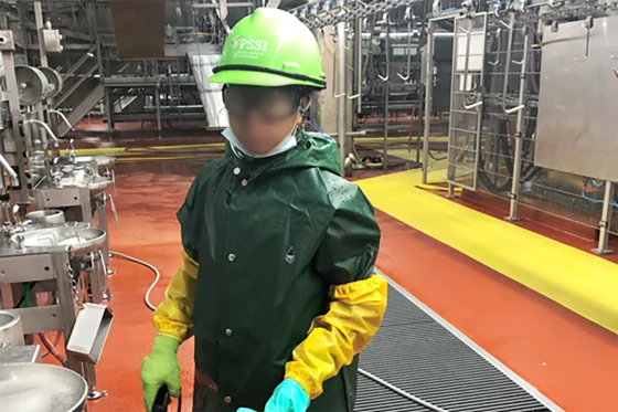 A child dressed in an industrial cleaning uniform - a lime green hardhat, a vinyl cleaning cloat, latex gloves. The background and location of the photo is in a meat packing facility. 